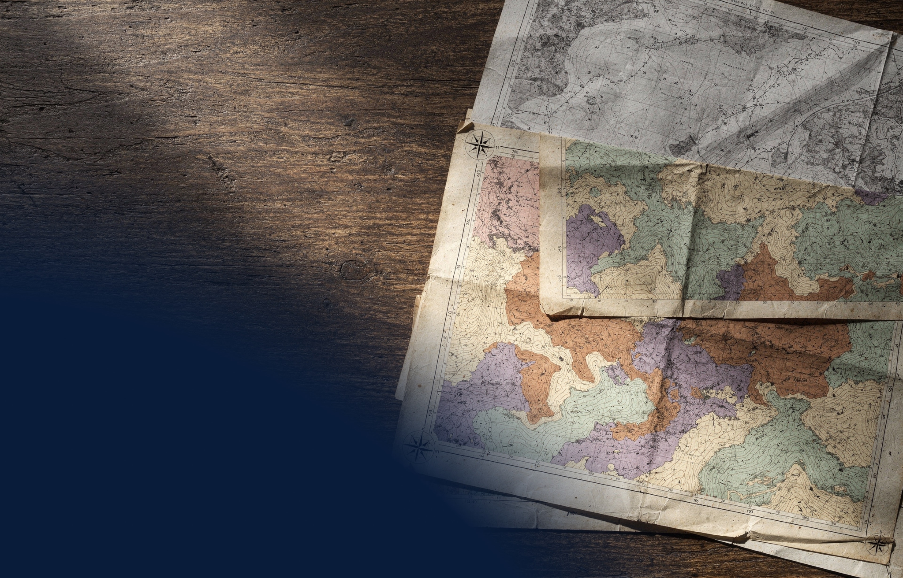 Overlapping geological and topographic maps on a weathered wooden desk — editorial overhead still life